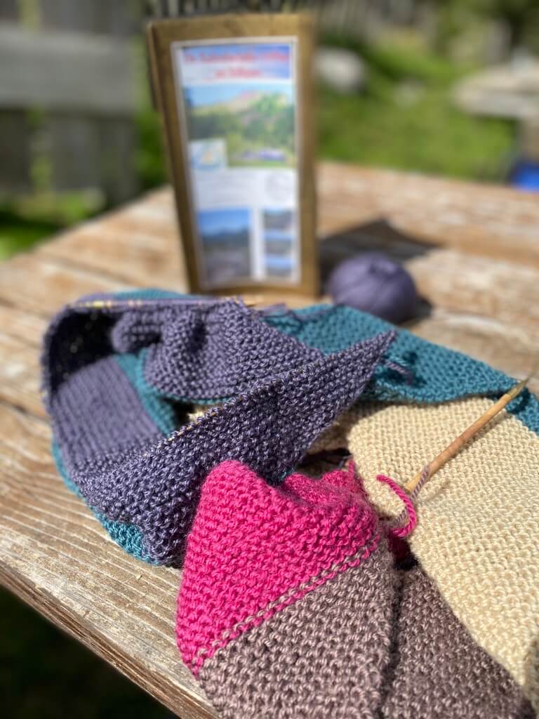 Ein angefangeness Strickstück liegt auf einem Holztisch, im Hintergrund ist eine Info-Karte mit Berglandschaft zu sehen