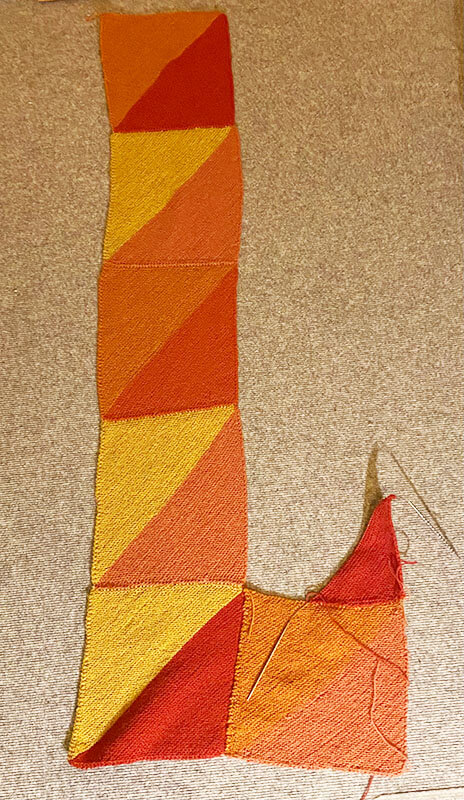 Der Fortschritt des Prism Blanket aus Purl Sohos "Good Wool" Unfertiges Strickstück ("UFO"): Der Anfang einer Decke aus diagonal gestrickten Quadraten in Rot- und Gelbtönen. Gezeigt wird eine fertige Reihe aus 5 Quadraten und eine Angefangene aus einem Quadrat und einem angefangenen Eck.
