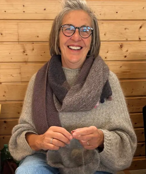 Brigitte Scherm sitzt, bekleidet mit einem Strickpulli und einem gestrickten Schal, vor einer holzvertäfelten Wand und strickt. Sie lacht in die Kamera.