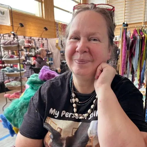 Portrait von Bettina Wagner im schwarzen T-Shirt, die Brille auf der Stirn lächslt sie verschmitzt in die Kamera. Im Hintergrund ein Wollmarkt.